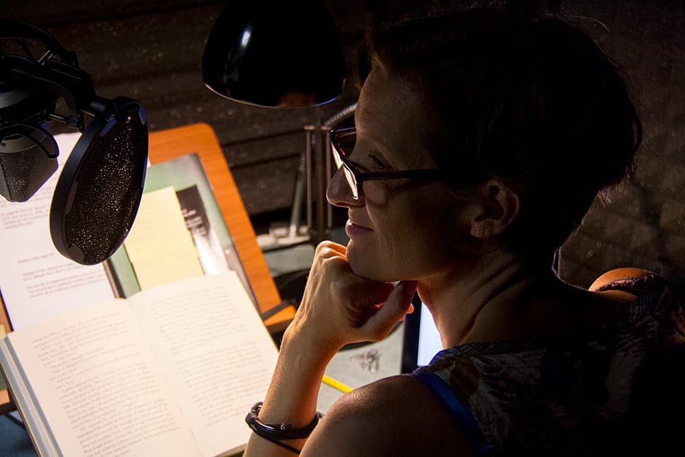 A professional voice-over artist recording an audiobook in a soundproof studio, with a microphone and book in front of her